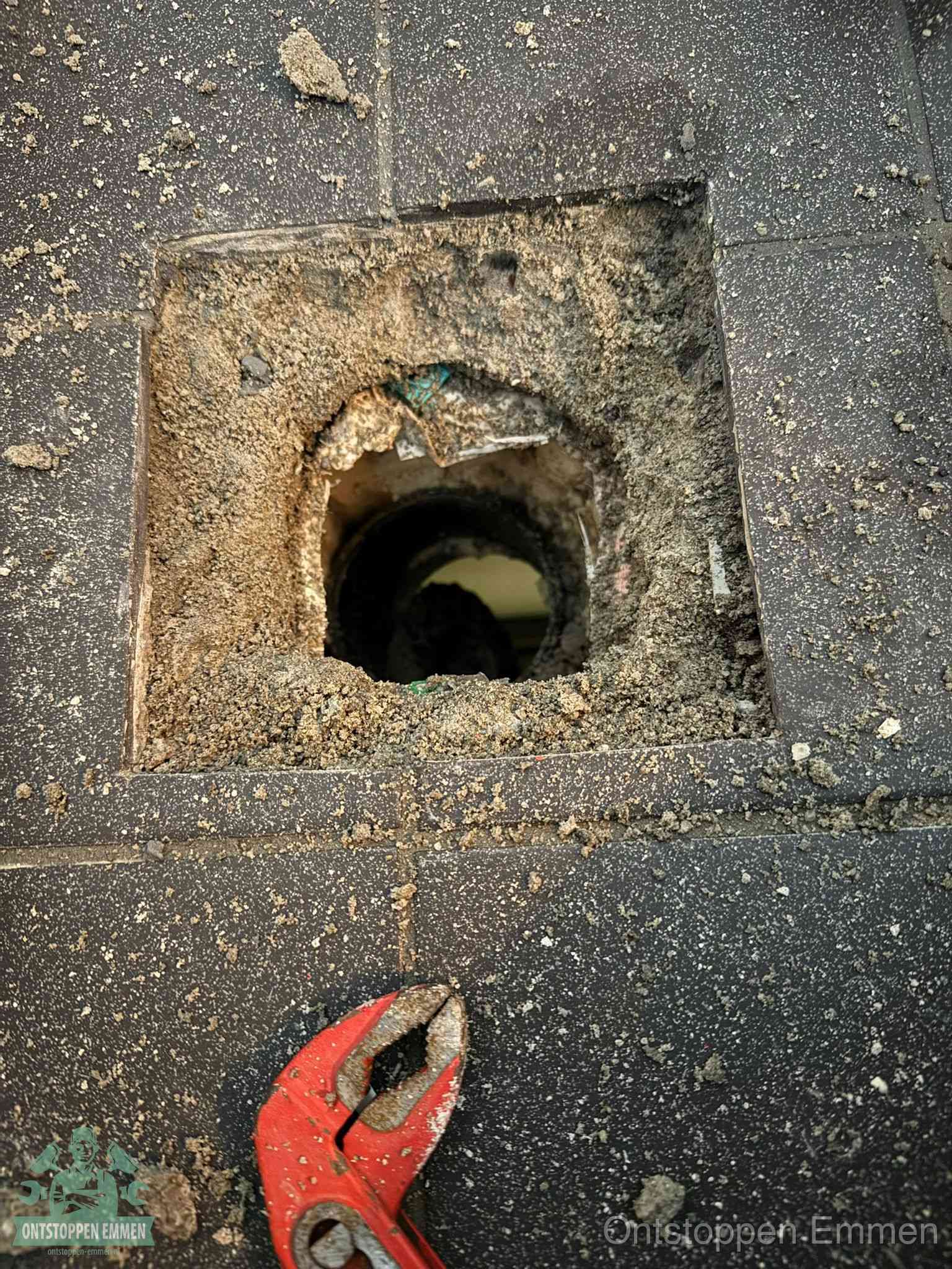 Een geopend rioolputje in een betontegelvloer met losgemaakt rooster en een zichtbare ronde afvoerbuis, het achterhalen waar de afvoerverstopping zit in Emmen