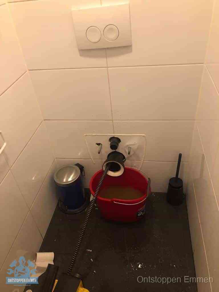 Een ontstopte wand-wc met verwijderde toiletpot waar een ontstoppingsveer in de wanduitgang verdwijnt, het achterhalen waar de verstopping zit in Emmen