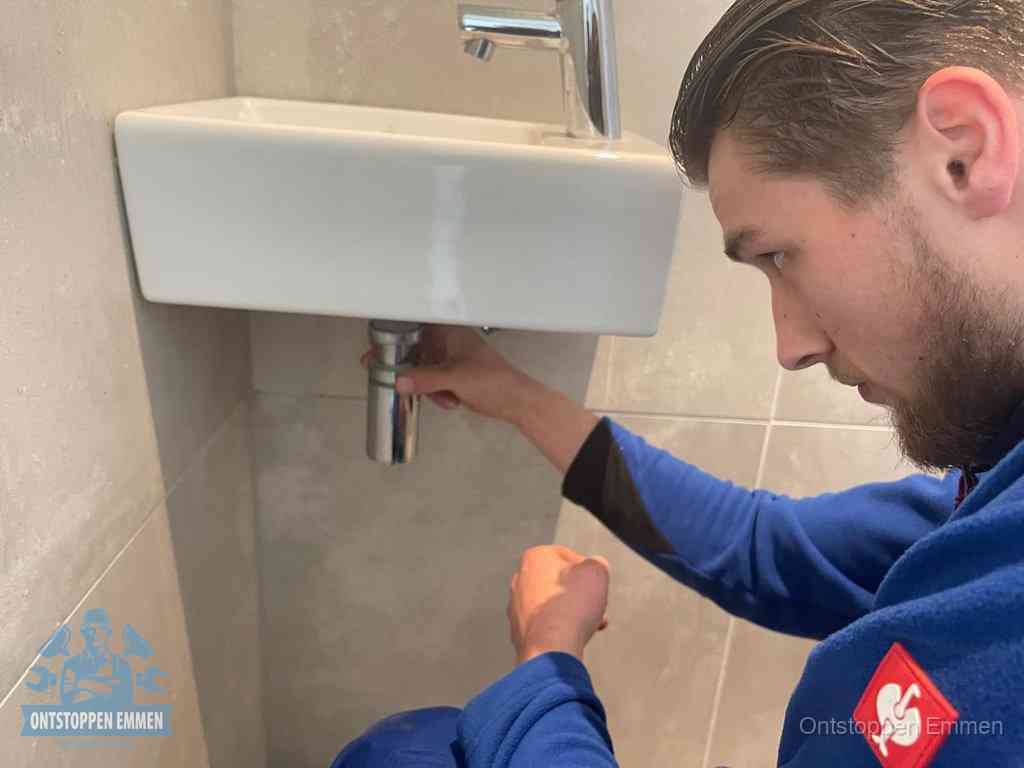 Een monteur draait met de hand de zwanenhalssifon onder een fonteinwasbak in de toiletruimte los om de bron van rioollucht in Emmen te vinden