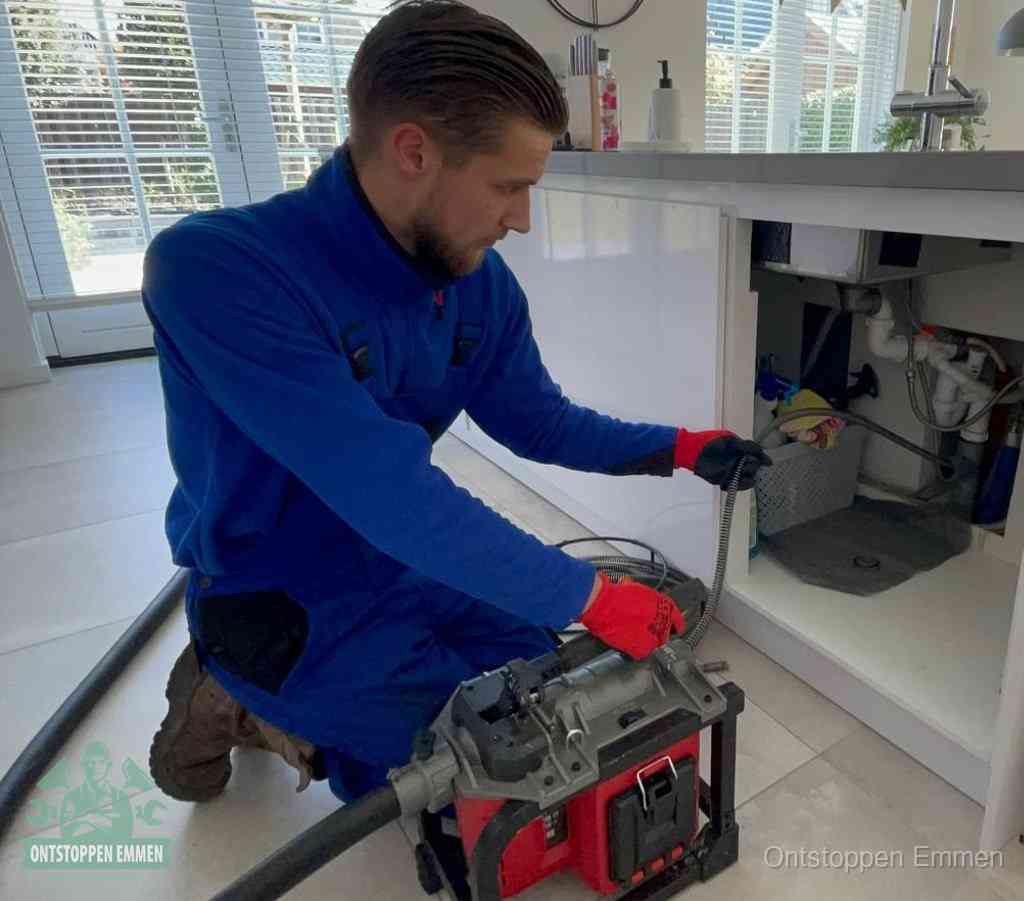 Een monteur met rode handschoenen knielt in een keuken en voert een elektrische ontstoppingsveer in tijdens het professioneel afvoer ontstoppen in Emmen